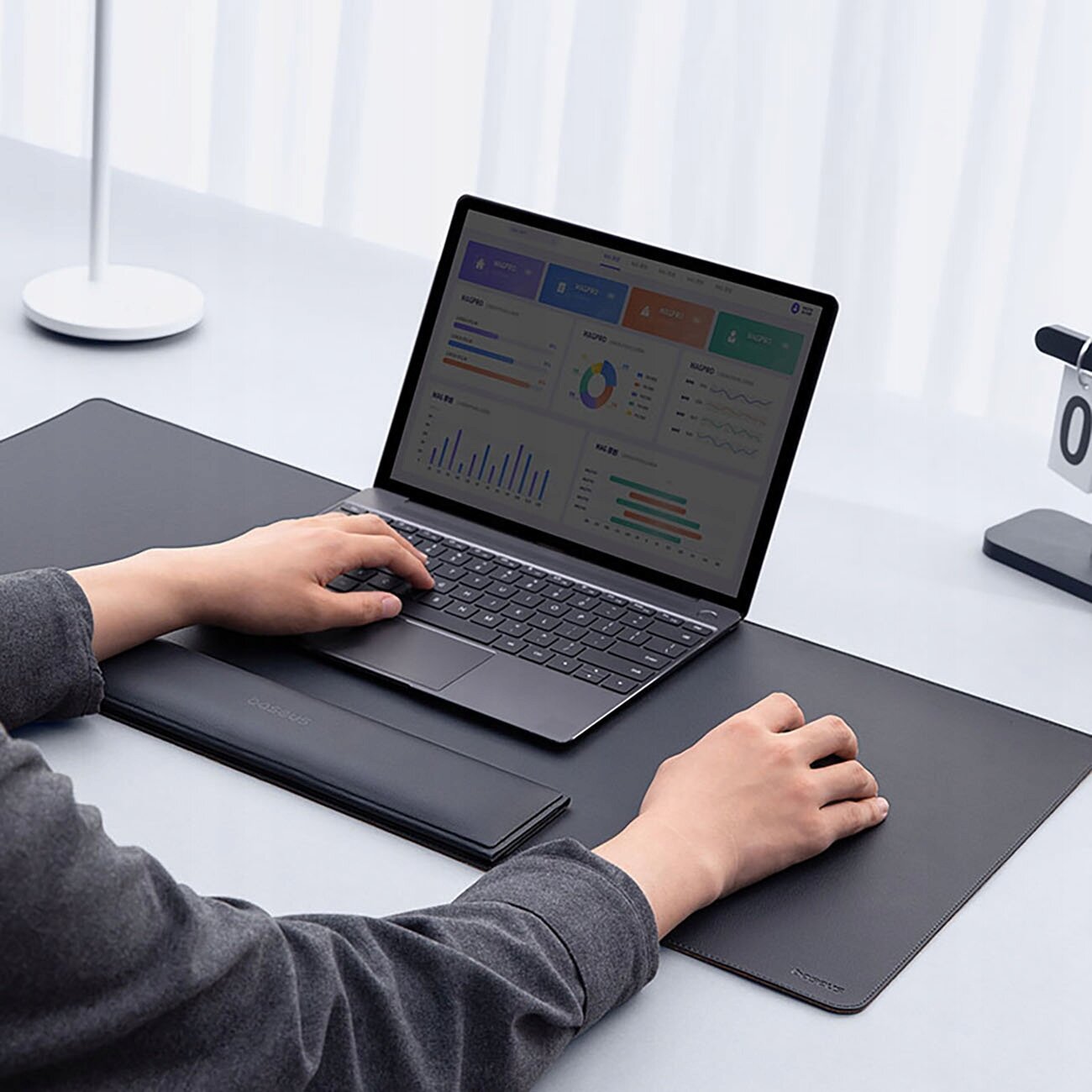 Osoba pracuje na laptopie umieszczonym na dużej, czarnej macie biurkowej z podpórką na nadgarstki. Na ekranie laptopa widoczne są wykresy i dane.