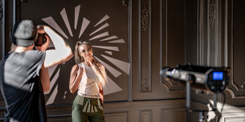 Fotograf wykonuje zdjęcie modelce w studiu; lampa z projekcją wzoru świetlnego tworzy dynamiczne tło na ścianie