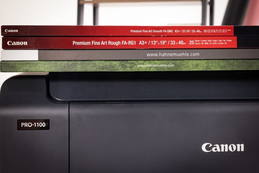 Drukarka CANON imagePROGRAF PRO-1100 Wi-Fi zbliżenie na górną część drukarki położone na niej opakowania z różnymi rodzajami papierów szeroki zakres obsługiwanych rodzajów i rozmiarów papieru dopasowanie papieru do zdjęcia ochrona przed zagnieceniem