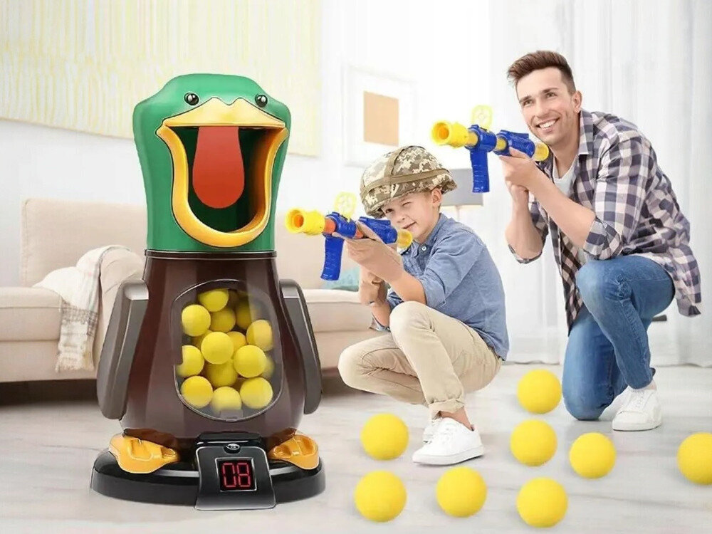 Gra plenerowa EXTRALINK Głodna kaczka Chłopiec w kasku wojskowym i dorosły mężczyzna celują z niebieskich pistoletów na piankowe kulki do interaktywnej zabawki w kształcie kaczki. Kaczka posiada przezroczysty brzuszek wypełniony żółtymi piłeczkami oraz cyfrowy licznik trafień u podstawy.