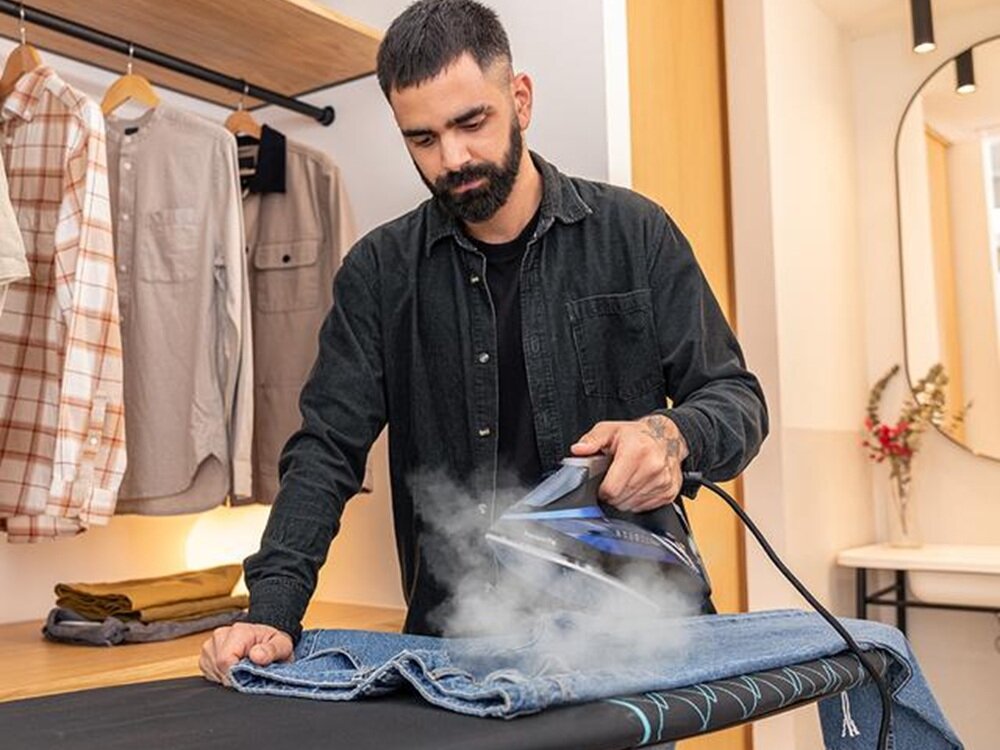 Osoba prasuje niebieskie dżinsy za pomocą żelazka, z którego wydobywa się para. W tle widać powieszone ubrania i półkę złożoną z kilku złożonych elementów odzieży.