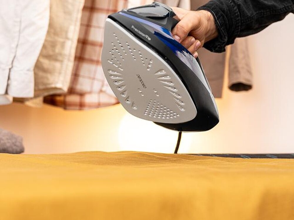 Osoba trzyma żelazko z widoczną stopą grzewczą z napisem \'CECOTEC\'. W tle widać powieszone ubrania.