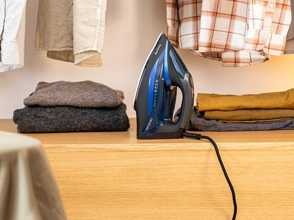Na drewnianej powierzchni stoi żelazko z niebieskimi i czarnymi elementami, obok którego leżą złożone ubrania. W tle widać wiszące koszule.