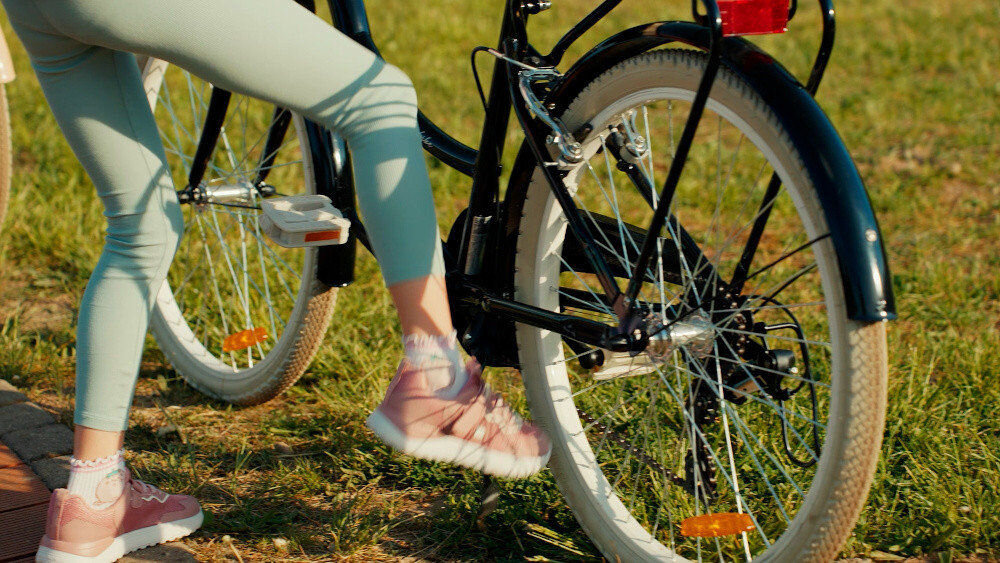 Rower młodzieżowy SUN BABY Flower Bike 24 cale dla dziewczynki Czarny zbliżenie na tylne koło osoba opiera nogę o stopkę w tle trawa stopka w centralnym punkcie ciężkości reflektory odblaskowe z przodu z tyłu i na szprychach roweru lepsza widoczność na drodze bezpieczeństwo dzwonek