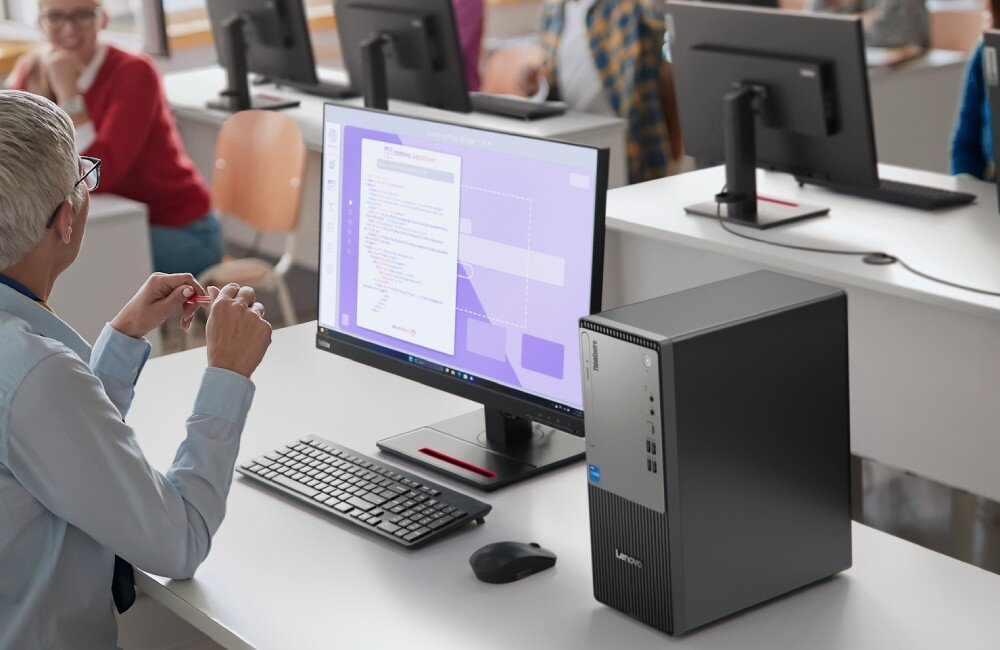 Komputer LENOVO ThinkCentre neo 50t Gen 5 Kobieta w okularach siedząca w jasnej sali lekcyjnej lub biurowej przodem do monitora z fioletowym interfejsem programu, obok którego na blacie biurka stoi czarna obudowa komputerowa typu tower, obudowa, wielkość, przeznaczenie