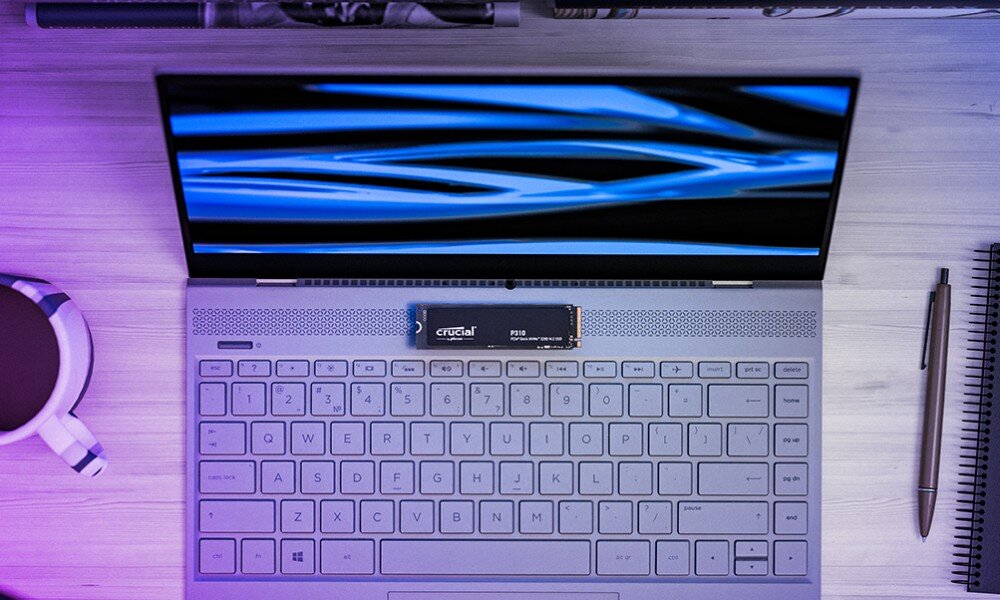 Dysk CRUCIAL P310 Widok z góry na otwartego laptopa z wyświetlaczem pokazującym niebieskie fale, dysk SSD 'crucial P310' położony na klawiaturze obok kubka i długopisu, energooszczędność