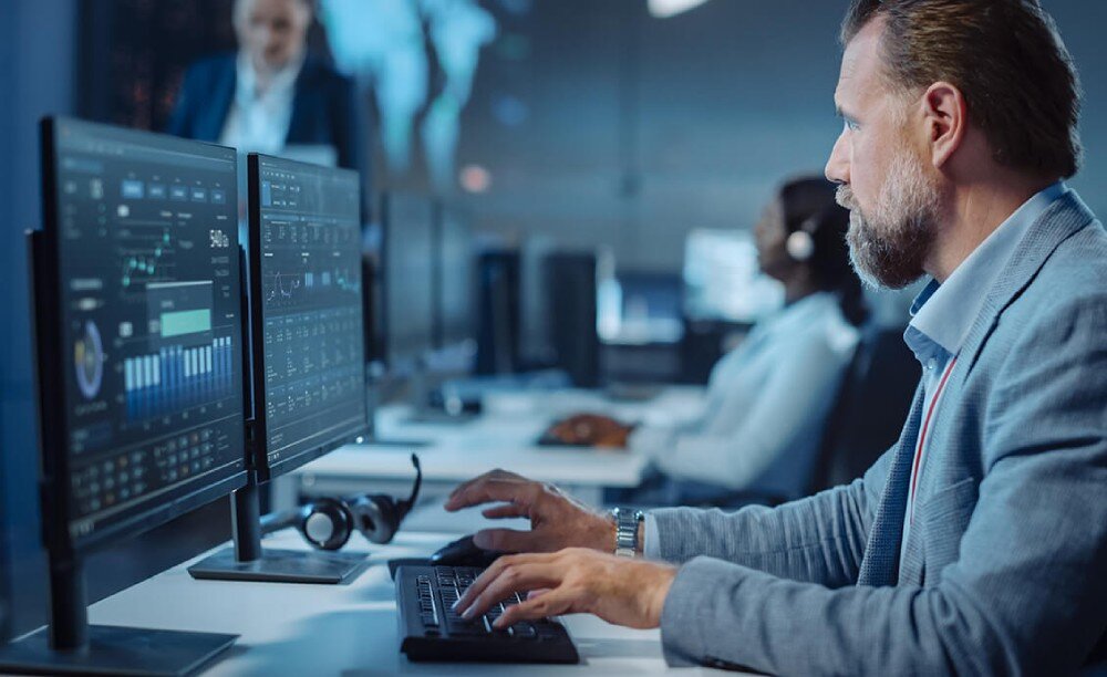 Procesor INTEL Core Ultra 7-265K Mężczyzna w garniturze pracujący przy dwóch monitorach z wykresami i danymi w biurze z innymi osobami na dalszym planie, zabezpieczenia, ochrona