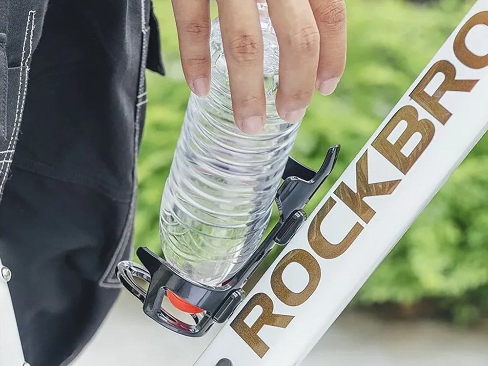 Osoba wkłada plastikową butelkę do czarnego koszyka na bidon zamontowanego na białej ramie roweru z napisem \'ROCKBROS\'.
