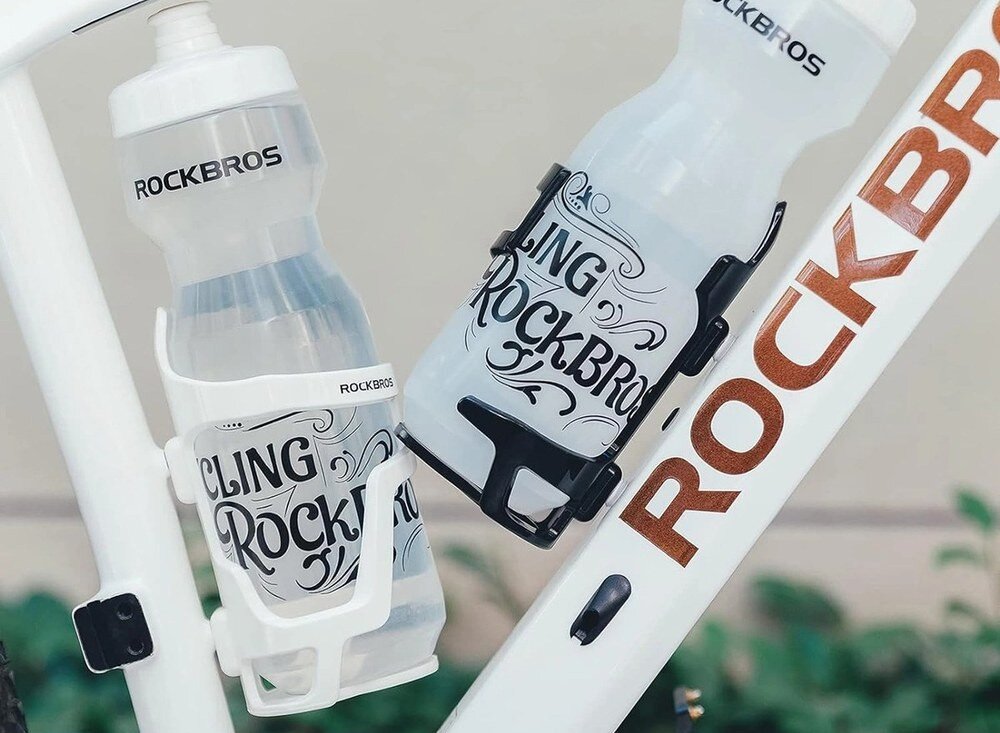 Koszyk na bidon ROCKBROS 31210012002 Dwa uchwyty Rockbros z bidonami zamocowane na białej ramie roweru, ujęcie z bliska na zielonym tle roślinności.