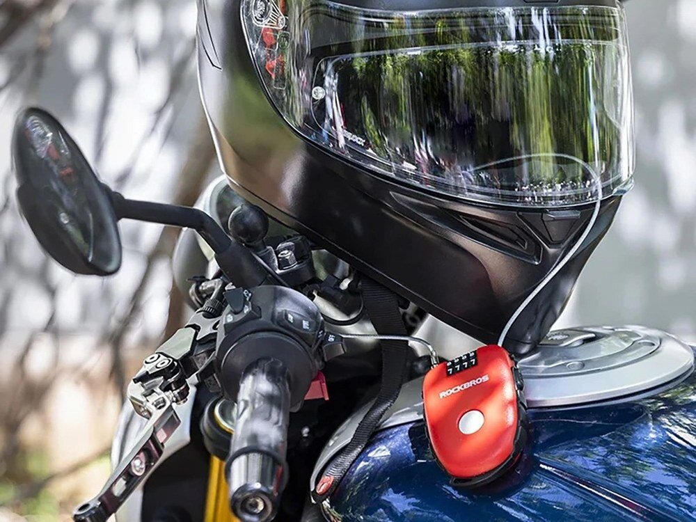 Na zdjęciu widoczny jest kask motocyklowy umieszczony na motocyklu, obok którego znajduje się czerwone zapięcie z napisem \'ROCKBROS\'.