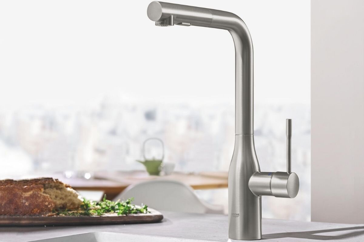BATERIA KUCHENNA GROHE ESSENCE 30270DC0 STAL SZCZOTKOWANA Bateria kuchenna ze stali nierdzewnej prezentowana na blacie w kuchni, z widokiem na stół z pieczywem