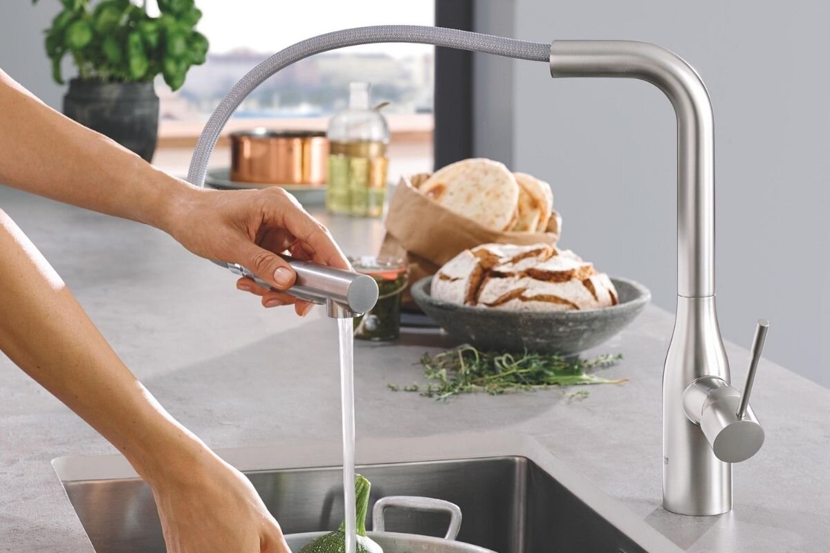 BATERIA KUCHENNA GROHE ESSENCE 30270DC0 STAL SZCZOTKOWANA Osoba używa wyciąganej wylewki stalowej baterii kuchennej, kierując strumień wody na warzywa w zlewie wylewka 360° wyciągana końcówka EasyDock dwa rodzaje strumienia klasyczny prysznicowy