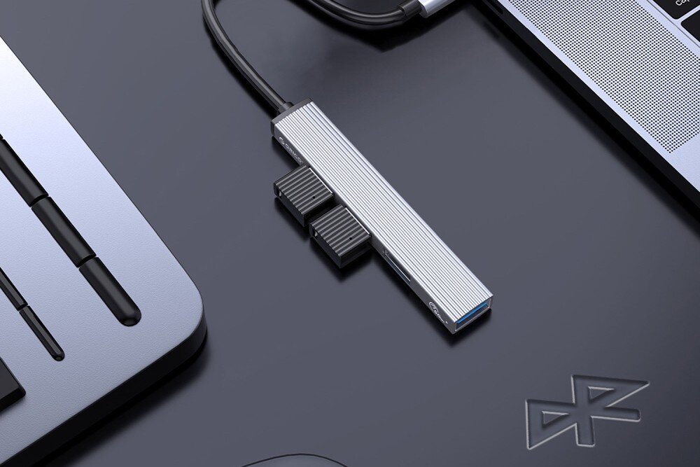 Hub ORICO AH-13-GY-EP-HW Srebrny adapter USB-C z podłączonymi dwoma pendrive'ami leży na czarnym biurku obok laptopa i innego urządzenia elektronicznego, opis