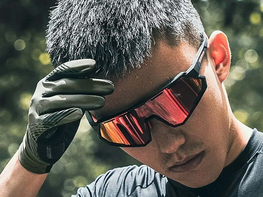 Mężczyzna w czarnych rękawiczkach Rockbros poprawia okulary rowerowe z czerwonymi szkłami, zielone tło.