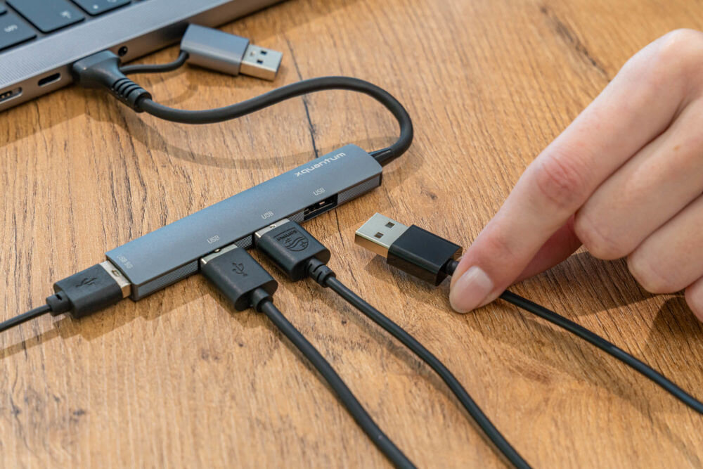 Hub USB XQUANTUM UH100 4w1 hub podpięty do komputera przez port USB-C obok 4 wtyczki USB-A ułożone obok portów adaptera