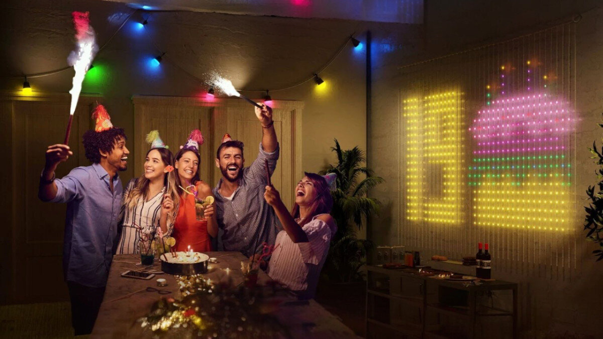 Kurtyna dekoracyjna GOVEE H70BC 400 LED Sieciowe 1.5 m Sieciowe Transparentny, kurtyna, światło, barwy, dekoracja, Wnętrze podczas imprezy urodzinowej – grupa przyjaciół świętuje przy stole, a na ścianie za nimi kurtyna LED tworzy świecący motyw tortu urodzinowego.