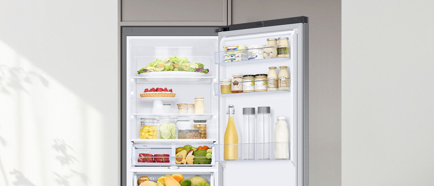 Otwarta lodówka z widocznymi półkami wypełnionymi różnorodnymi produktami spożywczymi, takimi jak owoce, warzywa, napoje i słoiki. Wnętrze jest jasno oświetlone, a drzwi lodówki zawierają dodatkowe półki na mniejsze produkty.