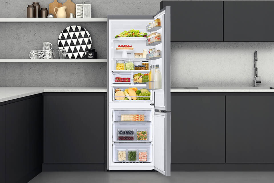 Otwarta lodówka w kuchni, wypełniona różnorodnymi produktami spożywczymi, takimi jak owoce, warzywa i napoje. W tle widoczne są ciemne szafki kuchenne i zlew.