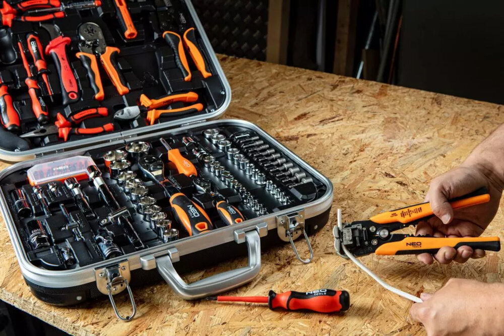 Zestaw narzędzi NEO TOOLS 01-310 otwarta walizka z zestawem na drewnianym blacie obok dłoń osoby ze szczypcami w dłoni szczypce 1000V wkrętaki precyzyjne ściągacze izolacji noże do kabli grzechotki nasadki adaptery bity