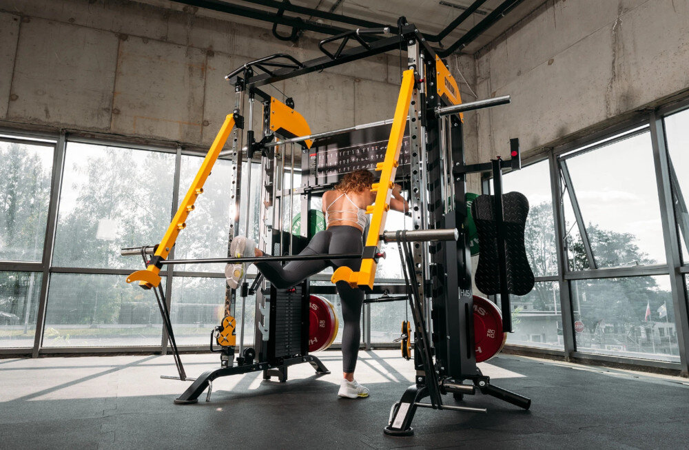 Suwnica HMS Cyklon X osoba ćwiczy nogi na suwnicy dwa stosy po 100 kg każdy z regulacją co 5 kg dopasowanie obciążenia do indywidualnych możliwości
