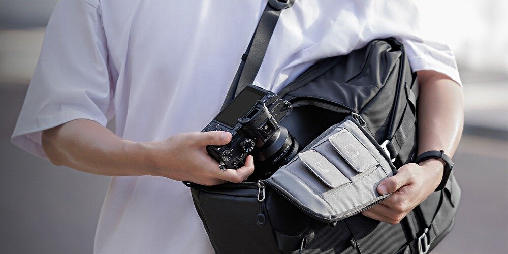 Otwarty plecak z boczną kieszenią na zamek; dłoń wyciąga aparat, obok metalowa klamra i karabińczyki