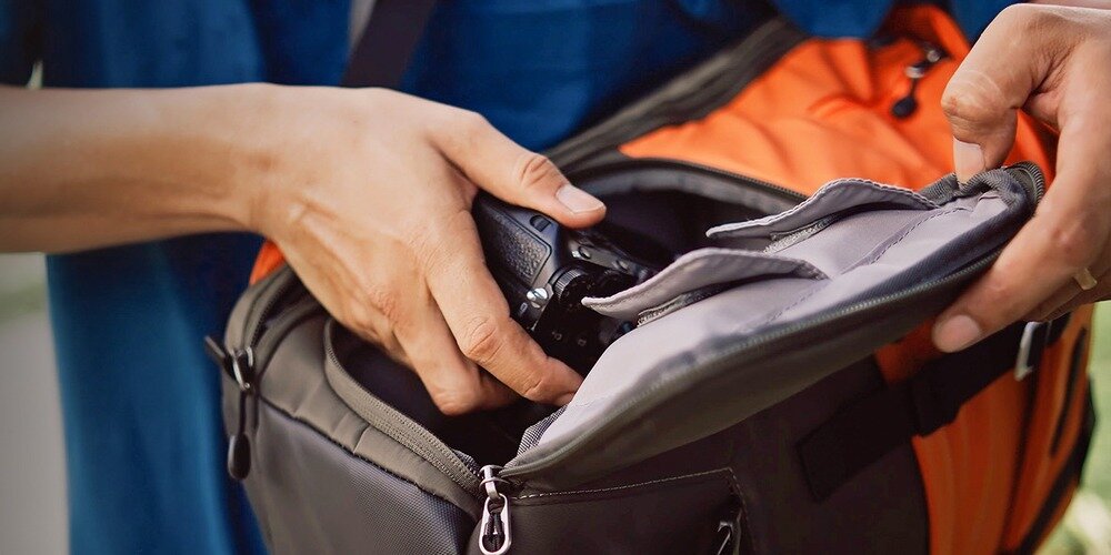 Otwarty plecak z boczną kieszenią na zamek; dłoń wyciąga aparat, obok metalowa klamra i karabińczyki