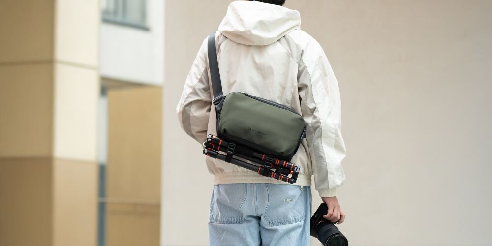 Młody mężczyzna stojący tyłem ma przewieszoną przez tułów torbę Alpha Sling Bag. Na spodzie torby przymocowany jest statyw