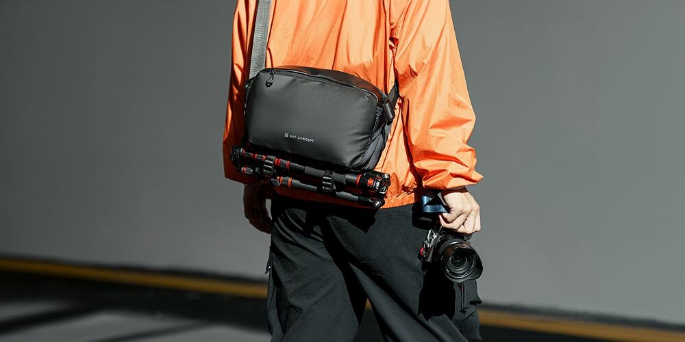 Młody mężczyzna stojący tyłem ma przewieszoną przez tułów torbę Alpha Sling Bag. Na spodzie torby przymocowany jest statyw