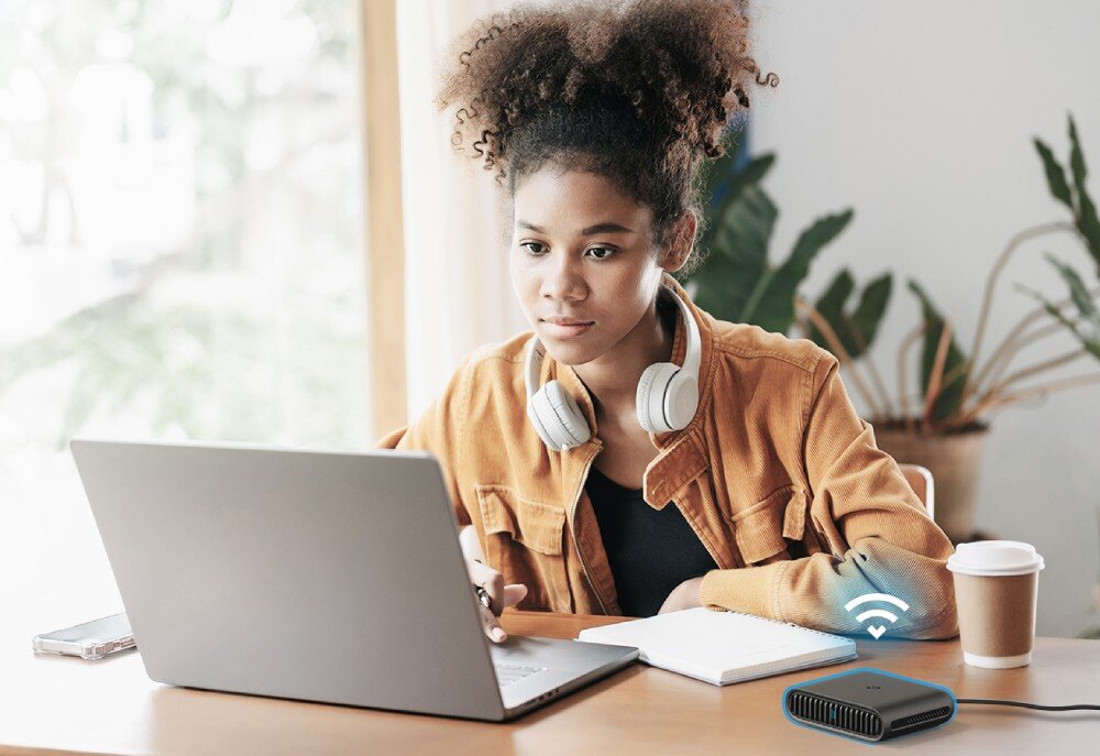Router TP-LINK TL-WR1502X Młoda kobieta w słuchawkach siedzi przy stole z laptopem i zeszytem. Obok znajduje się router z podświetlonym symbolem Wi-Fi. W tle duże okno i roślinność, tryby, działanie, modem, aplikacja
