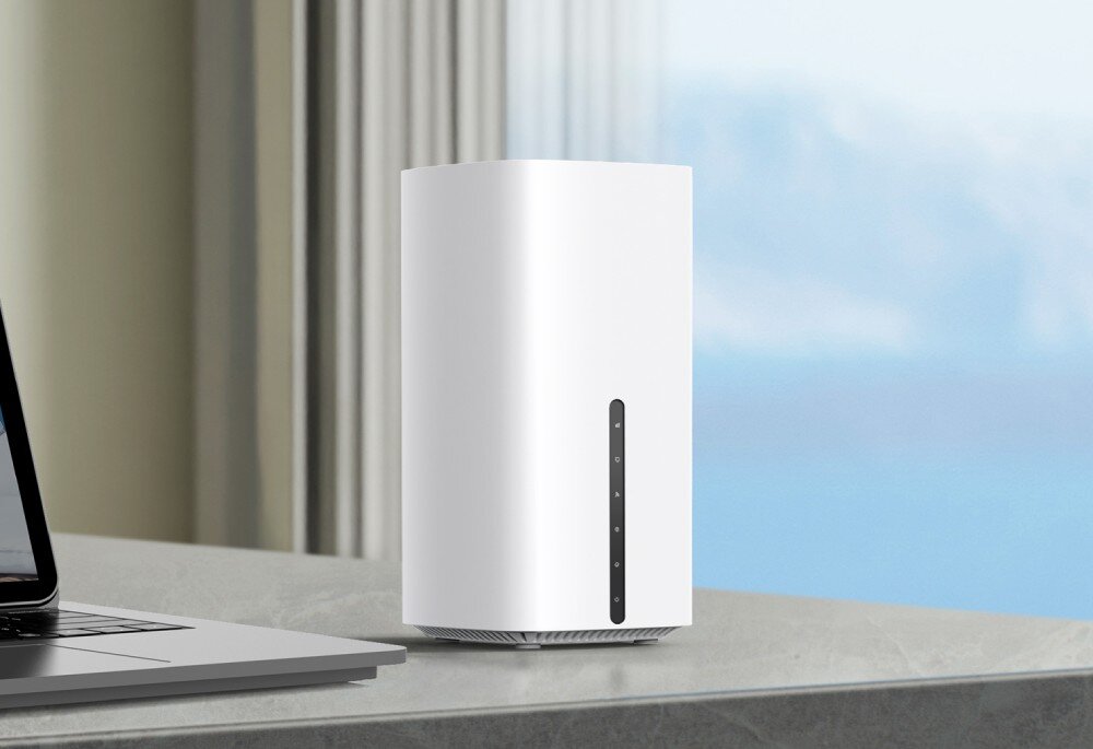 Router TP-LINK Archer NX600 5G Biały, pionowy router stoi na marmurowym blacie obok otwartego laptopa, w tle zasłona i rozmyty widok na zewnątrz, opis