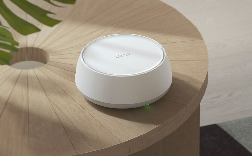 Router TP-LINK Deco BE25 Białe, cylindryczne urządzenie elektroniczne ustawione na drewnianym okrągłym stoliku. W tle widać fragment zielonej rośliny, działanie, sieć mesh, łączność Wi-Fi