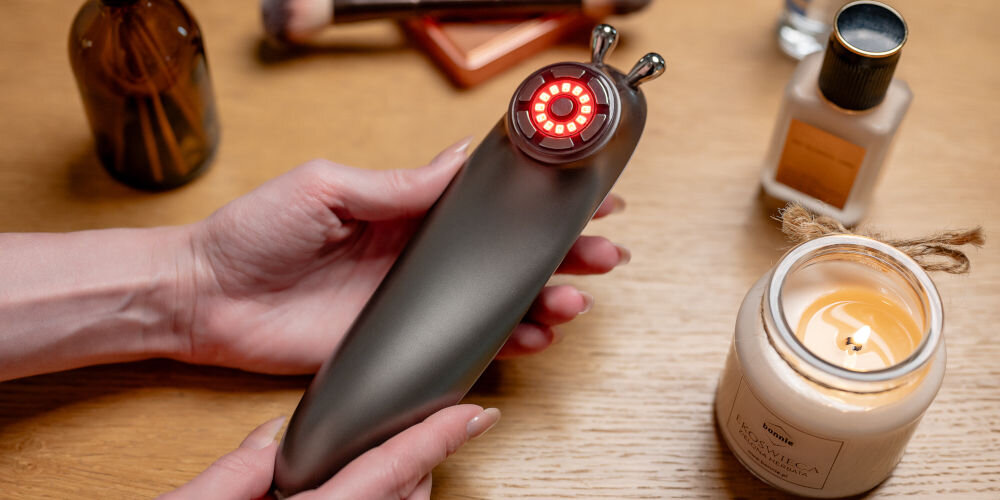 Urządzenie do liftingu twarzy HUMANAS Serenum srebrne urządzenie trzymane w rękach wydziela czerwone światło EMS Electrical Muscle Stimulation delikatne impulsy elektryczne delikatne bezbolesne skurcze naturalne sygnały nerwowe poprawia elastyczność do użytku domowego i podczas podróży