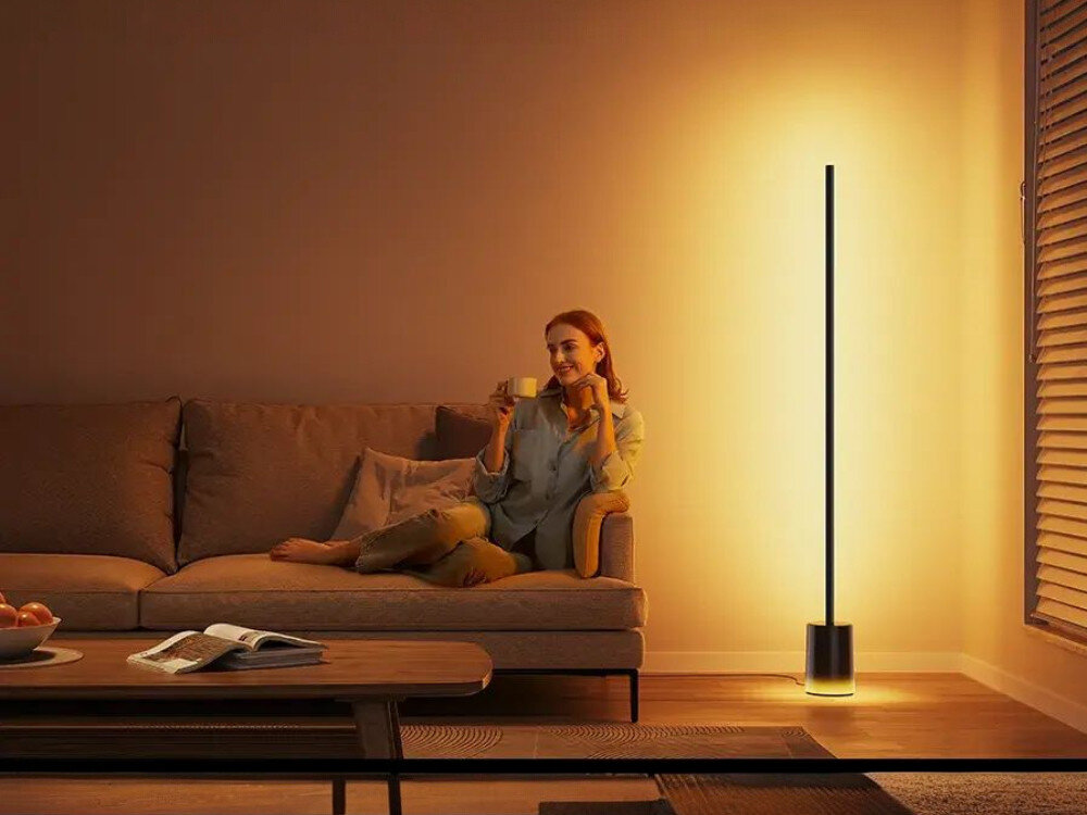 Lampa podłogowa GOVEE H607C, muzyka, rytm, scena, emocje, Relaksująca rudowłosa kobieta w szarej bluzce siedzi na brązowej sofie z kubkiem herbaty, obok smukła czarna lampa podłogowa Govee emitująca ciepłe żółte światło (ok. 2700K), oświetlająca nowoczesny salon z drewnianą podłogą, stolikiem z owocami i książkami.