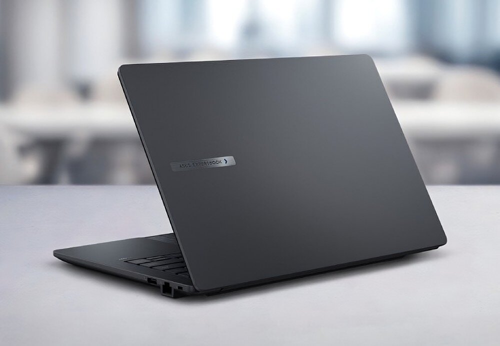 Laptop ASUS ExpertBook B1403CVA-S60475X Widok laptopa od tyłu, zamknięta obudowa w ciemnym kolorze z metalicznym logo producenta. Tło jest rozmyte i przedstawia biurowe wnętrze, pamięć RAM, dysk SSD, pojemność
