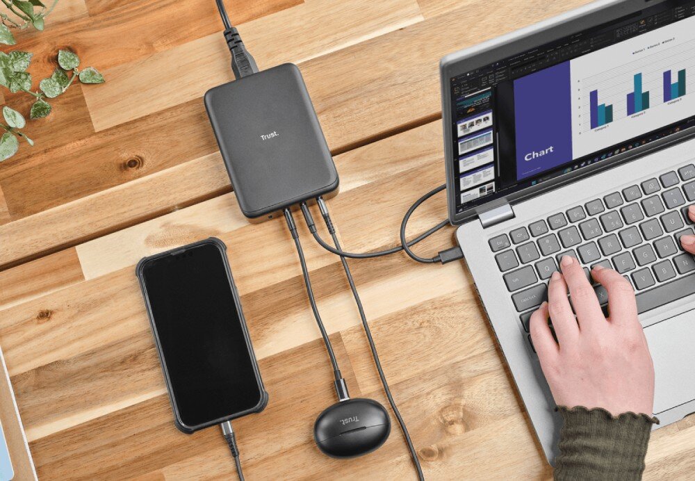 Zasilacz do laptopa TRUST Maxo 25413 240W Widok z góry na drewniane biurko z ładowarką zasilającą jednocześnie laptopa, telefon i słuchawki bezprzewodowe podczas gdy dłoń pisze na klawiaturze