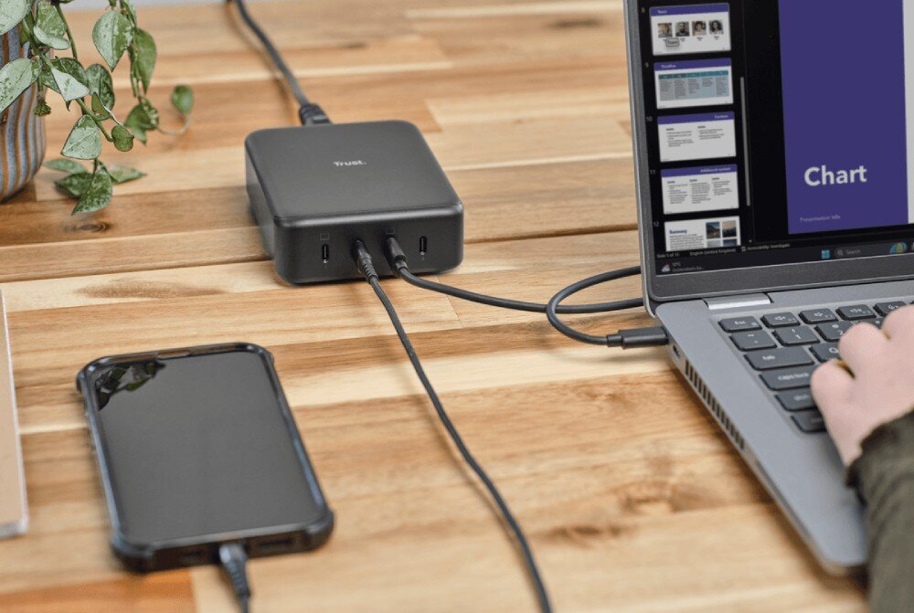 Zasilacz do laptopa TRUST Maxo 25413 240W Czarna ładowarka stacjonarna na drewnianym biurku podłączona przewodami do laptopa i smartfona z widoczną rośliną w tle
