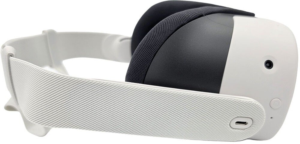 Na obrazie widoczne są gogle VR w kolorze białym z czarnymi elementami, posiadające teksturowany pasek i port USB-C. Na obudowie znajduje się okrągły przycisk oraz kamera.
