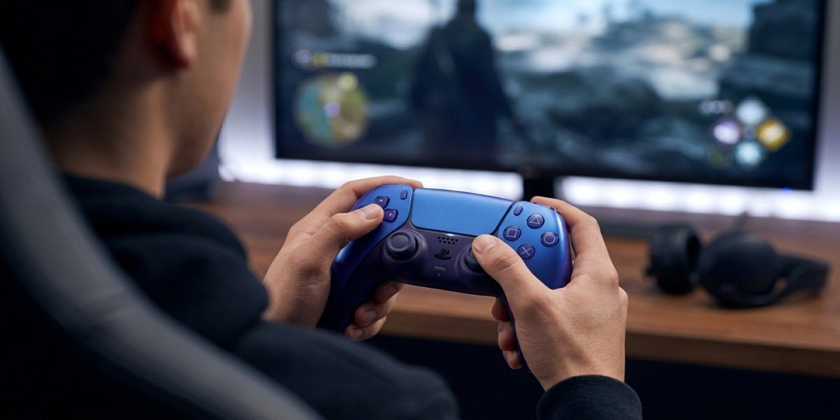 Osoba używająca niebieskiego kontrolera SONY DualSense, w tle widać monitor wyświetlający grę komputerową.