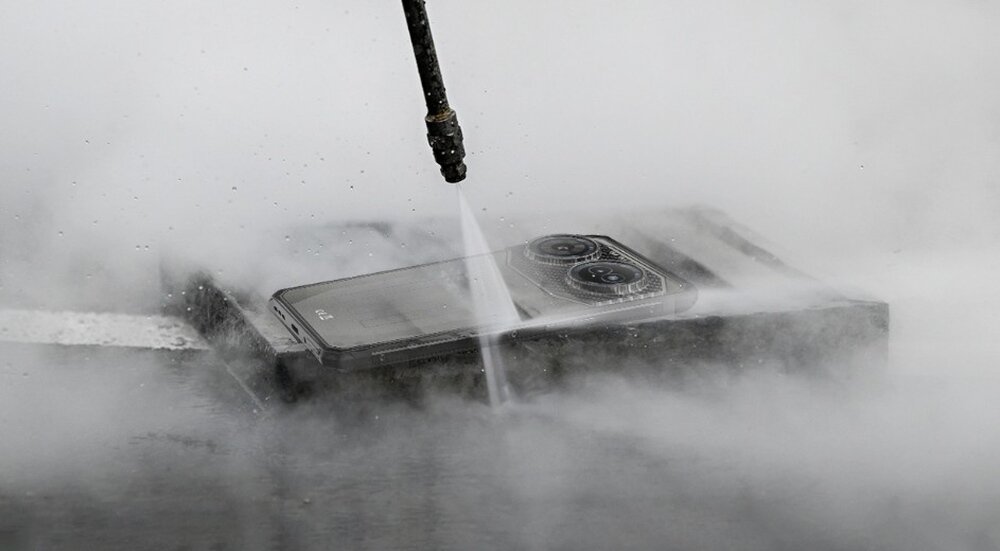 Smartfon jest poddawany działaniu strumienia wody pod ciśnieniem, co powoduje powstanie mgły wodnej wokół urządzenia. Na obudowie widoczne są dwa duże obiektywy aparatu.