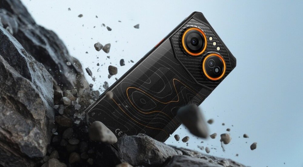 Smartfon z czarną obudową i pomarańczowymi akcentami leży na skałach, otoczony unoszącymi się drobinami kamieni. Na tylnej części urządzenia widoczne są dwa duże obiektywy aparatu oraz wzór przypominający mapę topograficzną.