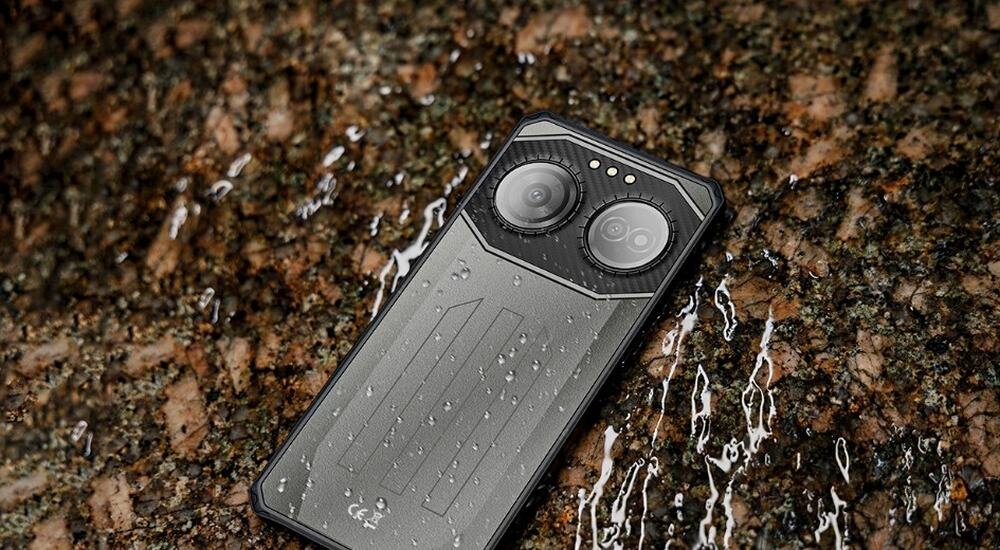 Smartfon z widocznymi dwoma dużymi obiektywami aparatu leży na mokrej powierzchni z kroplami wody. Obudowa urządzenia jest szara z geometrycznym wzorem.