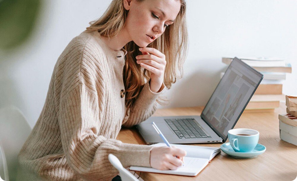 Router TP-LINK Deco BE25 Kobieta siedząca przy stole, notująca coś w zeszycie, korzystająca jednocześnie z laptopa. Na stole obok znajduje się filiżanka z napojem oraz kilka książek, obsługa VPN, rozwiązania