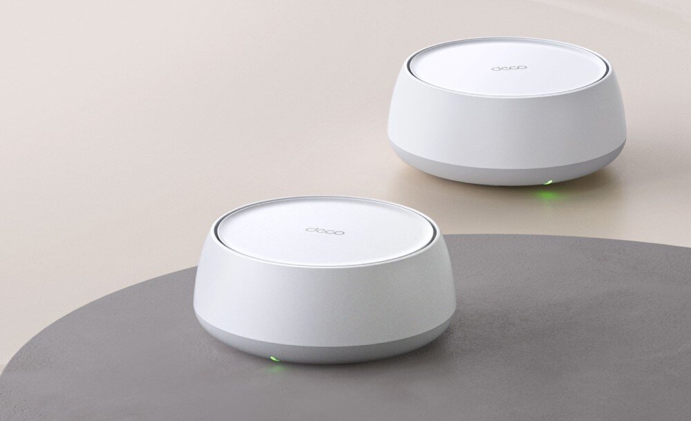 Router TP-LINK Deco BE25 Białe, cylindryczne urządzenie elektroniczne ustawione na drewnianym okrągłym stoliku. W tle widać fragment zielonej rośliny, działanie, sieć mesh, łączność Wi-Fi