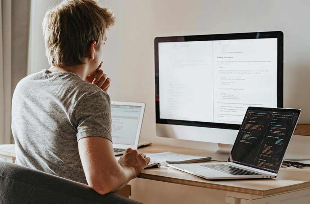 Router TP-LINK Deco BE25 Mężczyzna siedzący przy biurku, patrzący na ekran monitora. Przed nim znajduje się laptop, a na ekranach widoczne są fragmenty kodu programistycznego lub tekstu, ochrona sieci, wykrywanie zagrożeń