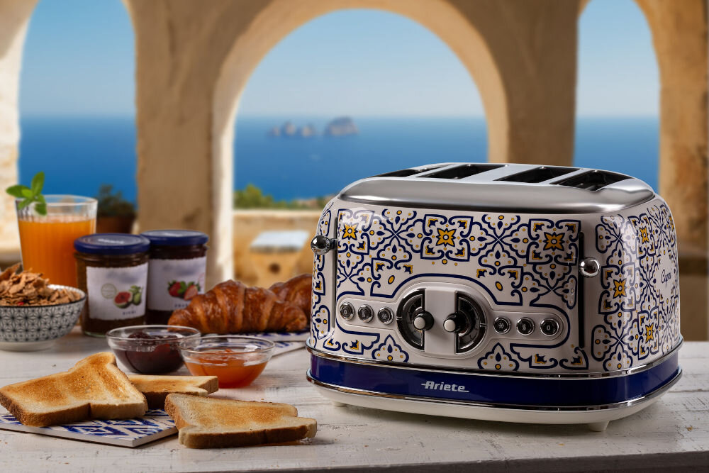 Toster ARIETE Capri 1560C optymalna chrupkość toster stoi na stole wzorzysta obudowa przyciski suwak otwory na stole rogale tosty dodatki w tle morze