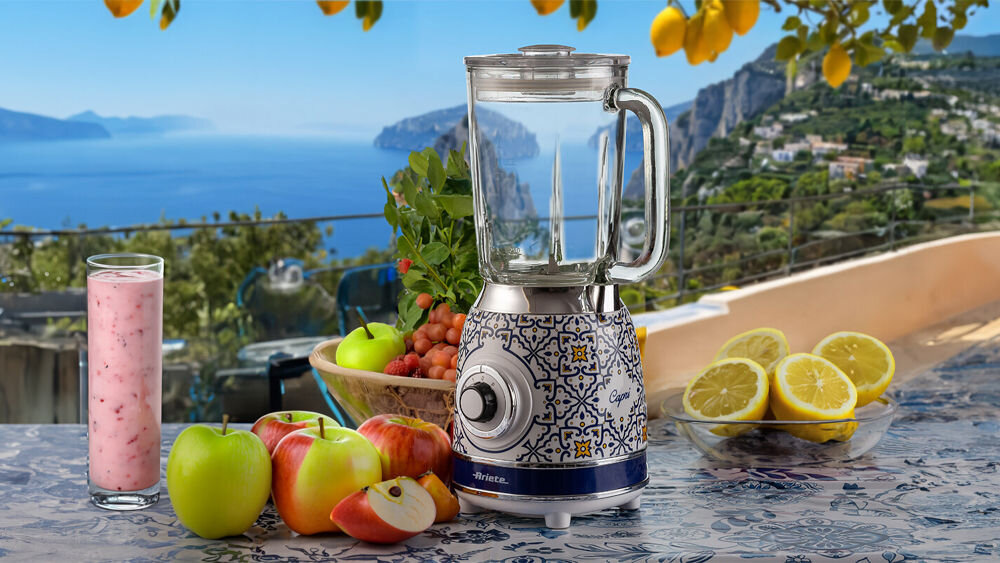 Blender kielichowy ARIETE 583 Capri blender stoi na blacie biało niebieska wzorzysta obudowa pokrętło przeźroczysty dzbanek różowy koktajl w szklance owoce w koszyku widok morza kuchenny kompan