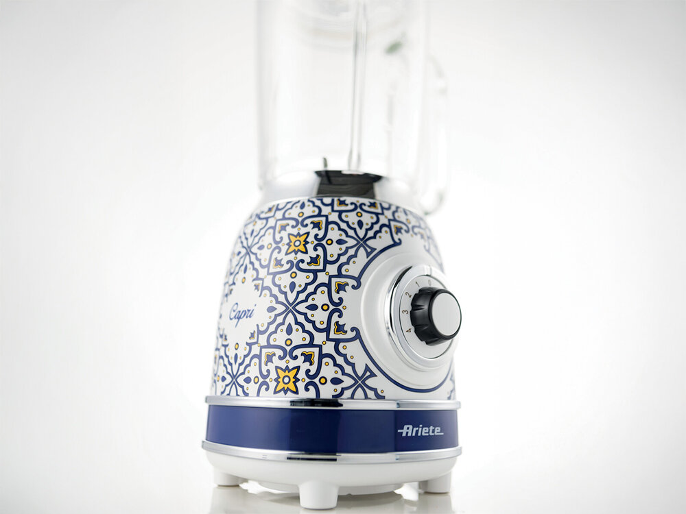 Blender kielichowy ARIETE 583 Capri białe tło blender pokrętło biało niebieska wzorzysta obudowa nóżki przeźroczysty kielich funkcjonalność w stylowym wydaniu Szklany kielich o pojemności 1,5 l 4 ostrza ze stali nierdzewnej silnik o mocy 1000 W smoothie aksamitne zupy koktajle mleczny 21,5 cm średnicy 18,5 cm długości 40,5 cm szerokości Biała obudowa niebieskie wzory żółte akcenty śródziemnomorska atmosfera