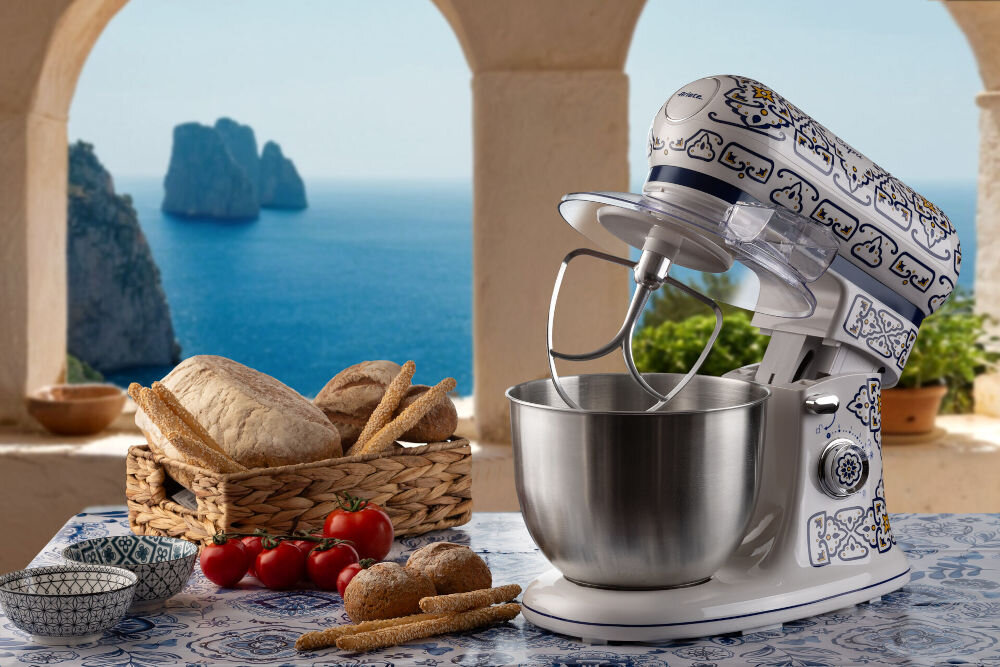 Robot kuchenny planetarny ARIETE 1588 Capri 2400W kuchenny pomocnik widok morza robot stoi na blacie otwarta pozycja mieszadło osłonka kolorowa obudowa biało niebieska elementy żółtego obok stoi pieczywo w koszyku i warzywa na blacie