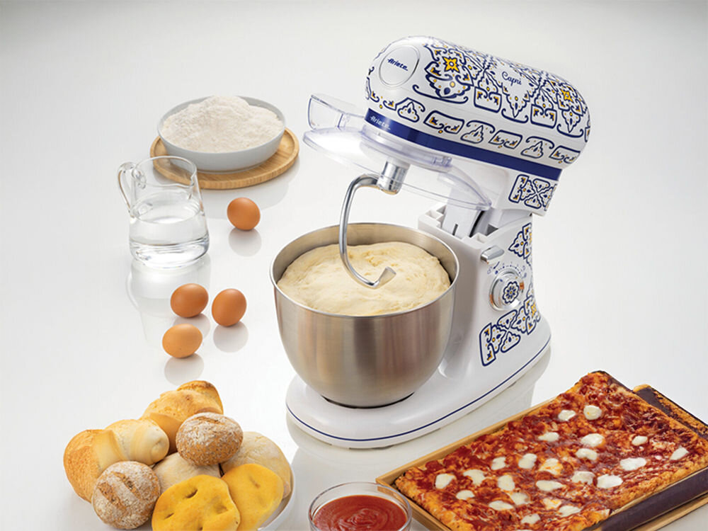 Robot kuchenny planetarny ARIETE 1588 Capri 2400W białe tło robot wzorzysta obudowa hak masa w misie wypieki i składniki obok urządzenia potężny silnik specjalny hak do zagniatania wyrabianie ciasta drożdżowego chlebowego na pizzę optymalne rezultaty oszczędza czas i energię