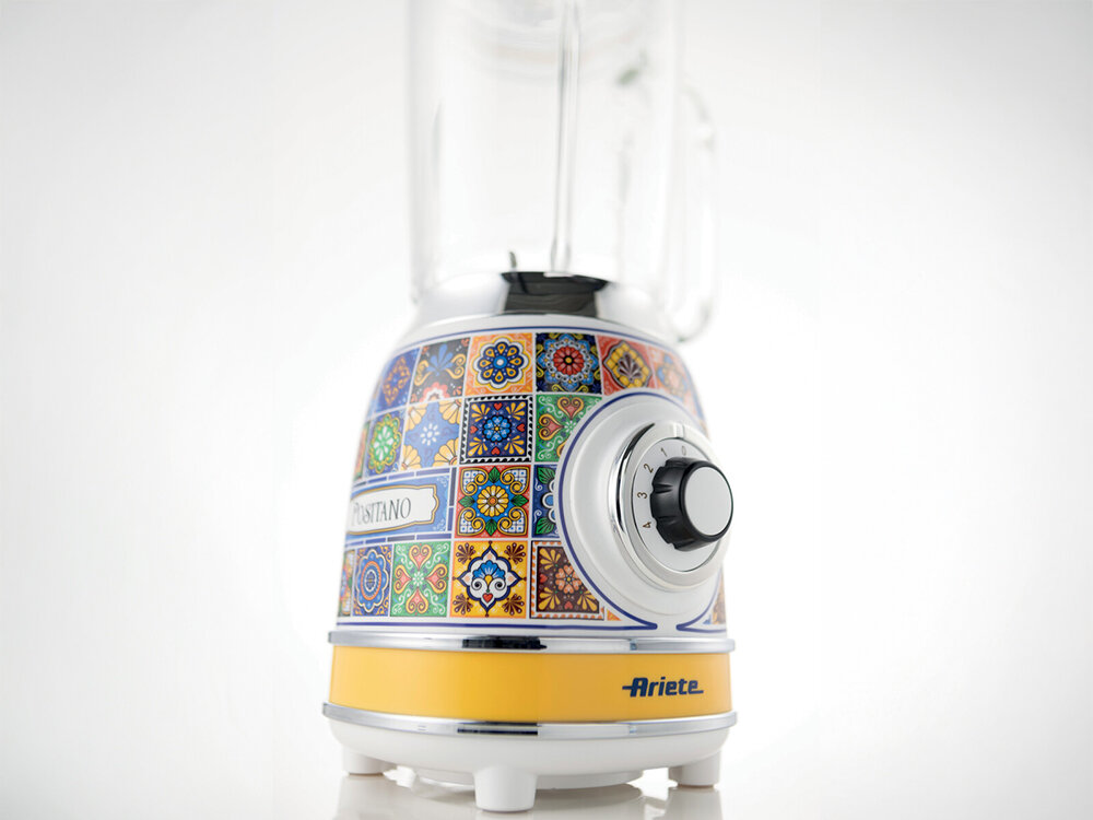 Blender kielichowy ARIETE 583 Positano białe tło blender pokrętło żółto niebieska wzorzysta obudowa nóżki przeźroczysty kielich funkcjonalność w stylowym wydaniu Szklany kielich o pojemności 1,5 l 4 ostrza ze stali nierdzewnej silnik o mocy 1000 W smoothie aksamitne zupy koktajle mleczny 21,5 cm średnicy 18,5 cm długości 40,5 cm szerokości Żółta obudowa mozaika śródziemnomorska atmosfera